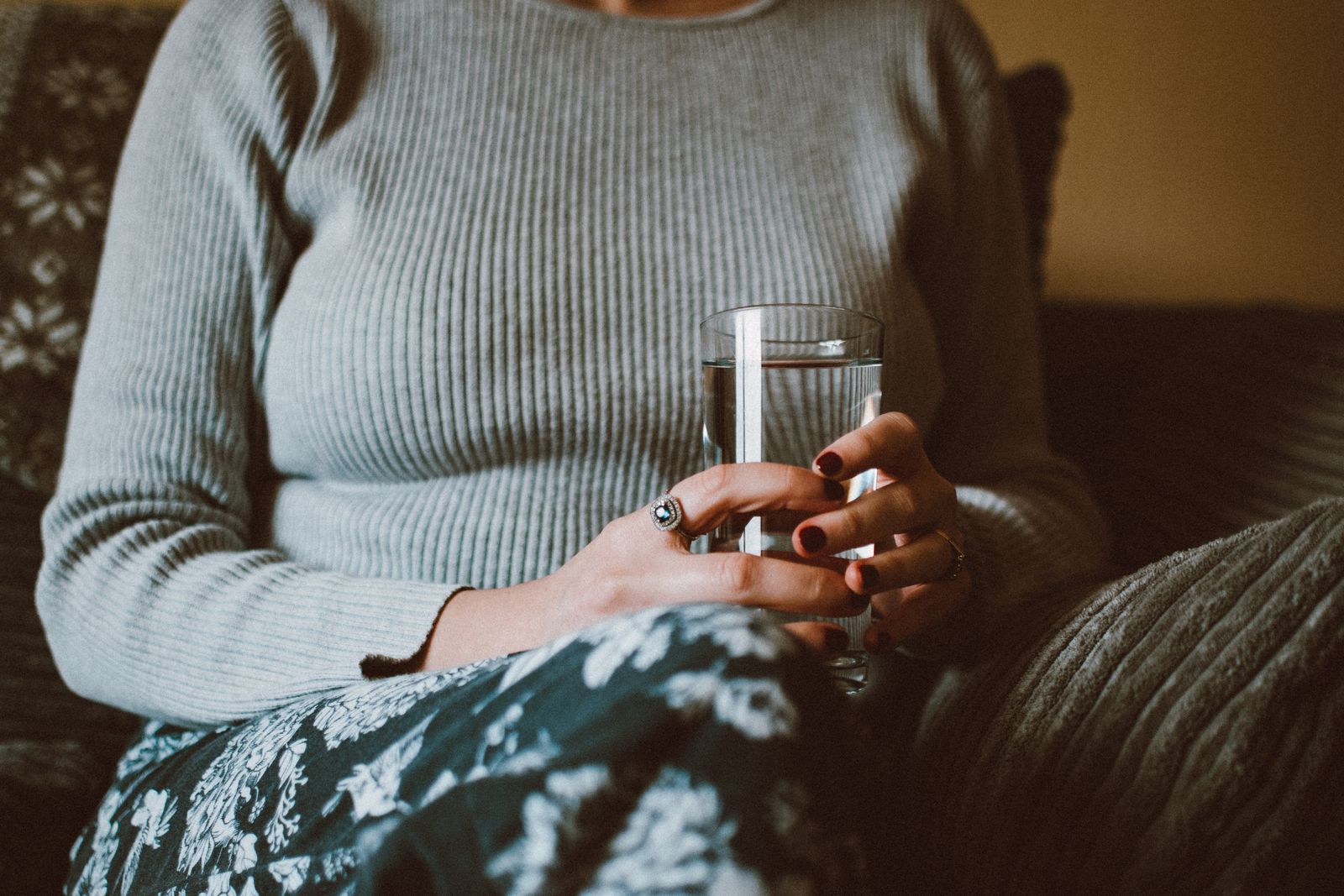 Mujer con jersey gris sentada en el sof&aacute; sosteniendo un vaso de agua con las dos manos