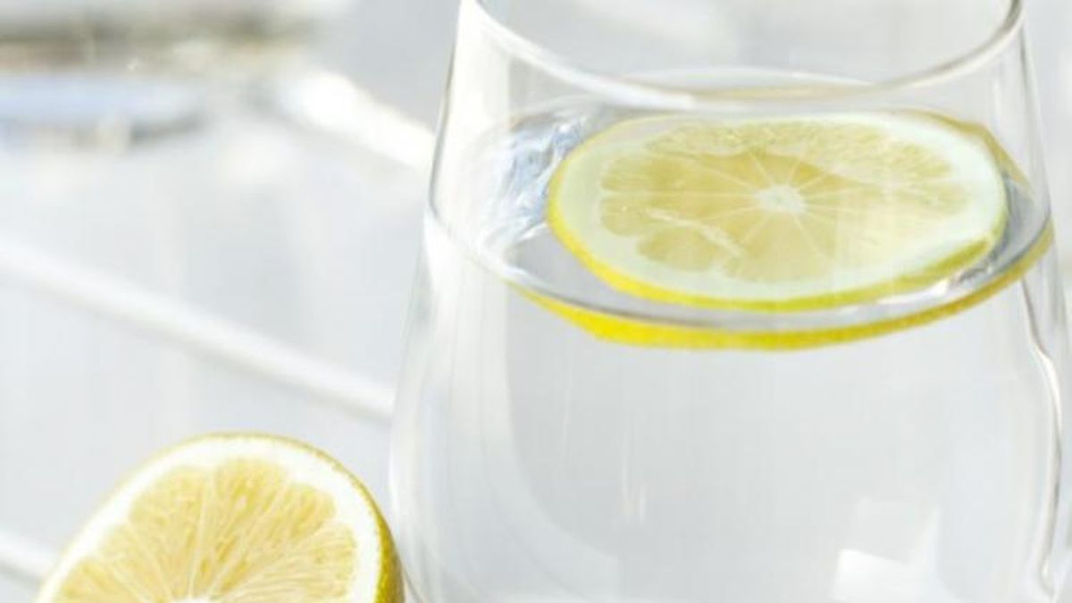 Vaso de agua transparente con una rodaja de lim&oacute;n flotando y medio lim&oacute;n al lado, sobre una mesa clara
