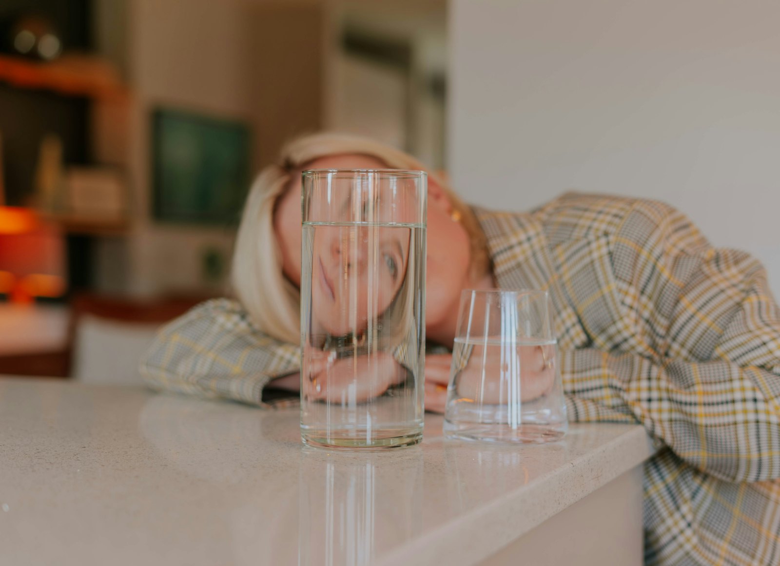Mujer cansada apoyando la cabeza sobre la encimera junto a dos vasos de agua, ilustrando el dolor de cabeza por deshidrataci&oacute;n