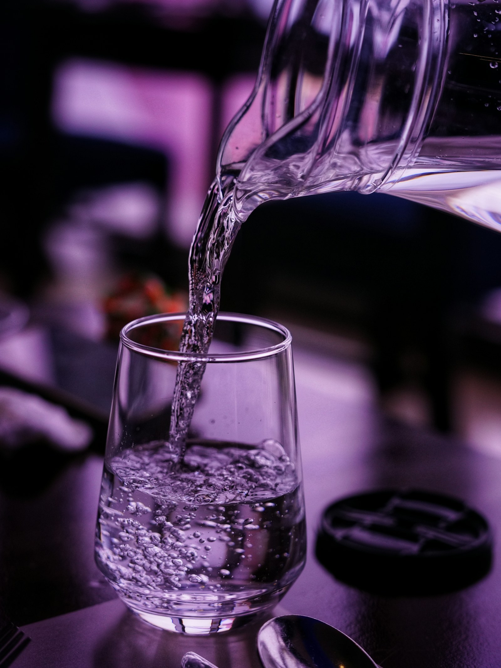 Jarra de cristal sirviendo agua en un vaso sobre una mesa puesta de restaurante, junto a los cubiertos