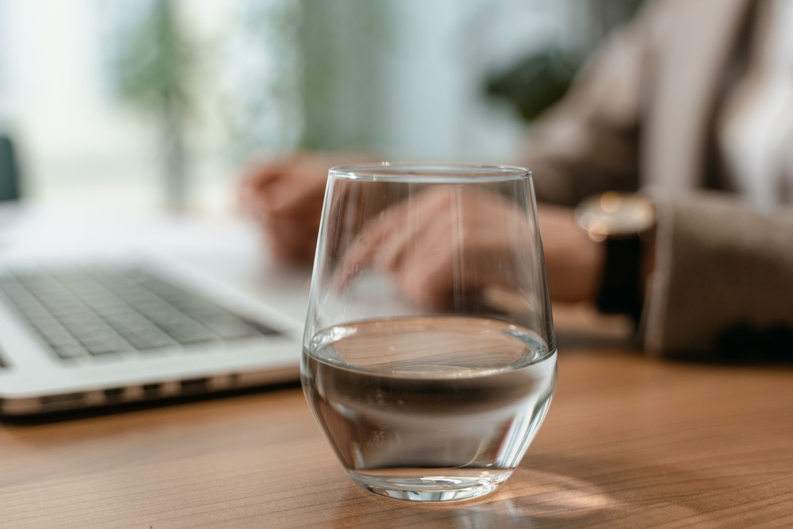 Vaso de agua sobre una mesa de oficina de madera, con un port&aacute;til y una persona escribiendo en el teclado desenfocada al fondo