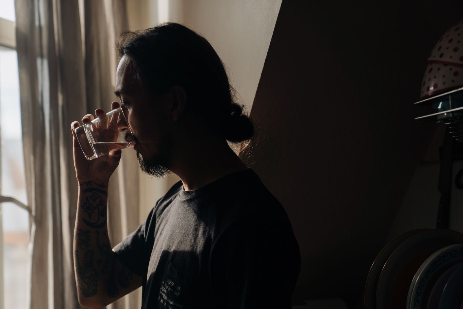 Hombre joven bebiendo un vaso de agua junto a una ventana con cortinas a primera hora de la ma&ntilde;ana
