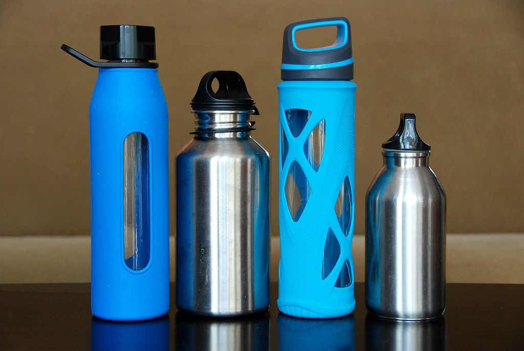 Cuatro botellas reutilizables de vidrio con funda de silicona y de acero inoxidable, alineadas sobre una superficie oscura
