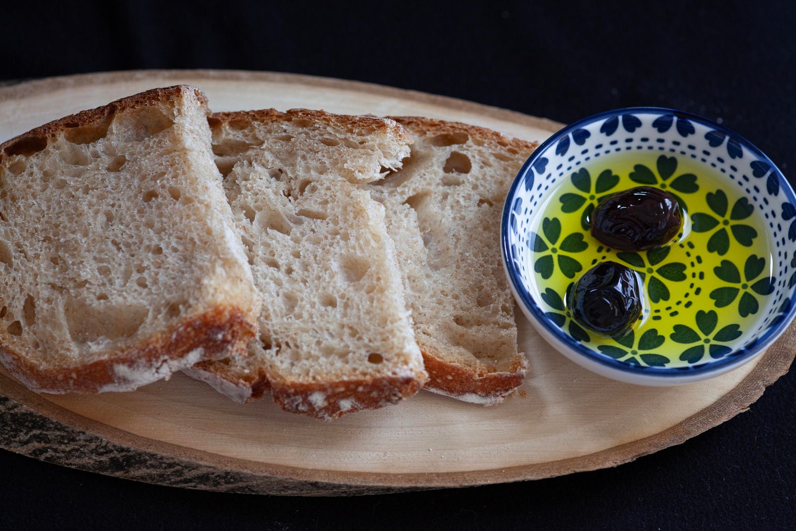 Rebanadas de pan de masa madre sobre una tabla de madera junto a un cuenco de cer&aacute;mica con aceite de oliva virgen extra y dos aceitunas negras