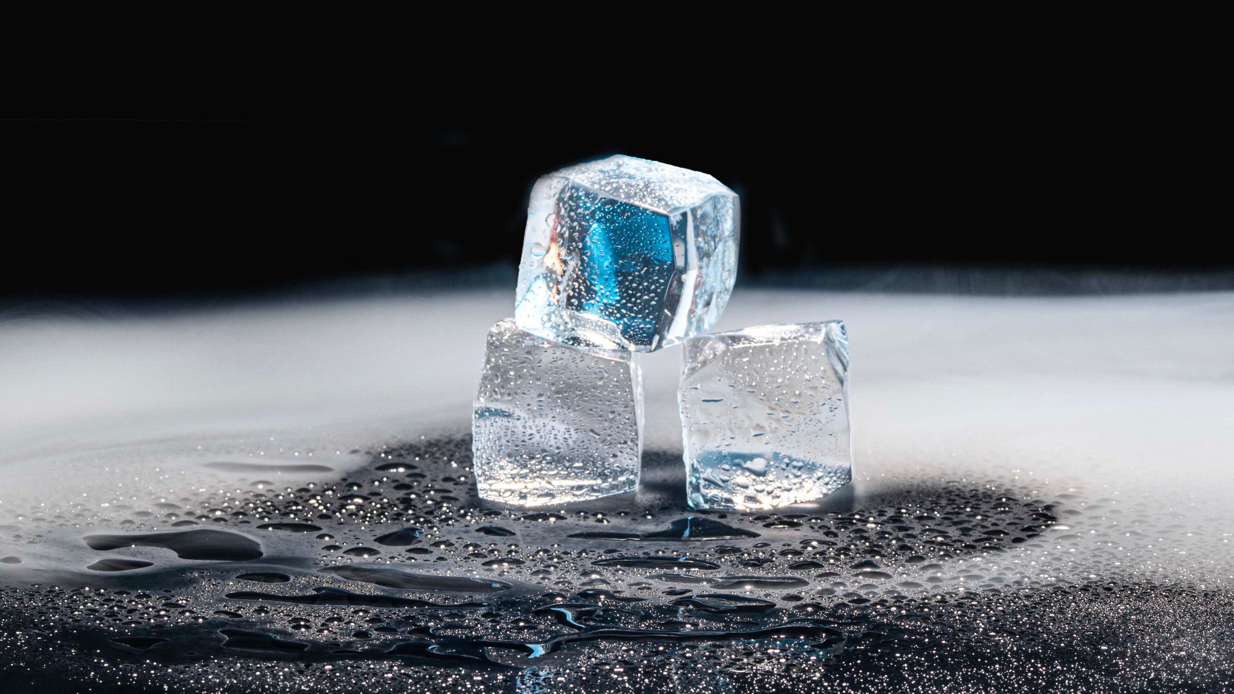 Tres cubitos de hielo transparentes derritiendose sobre una superficie oscura y mojada