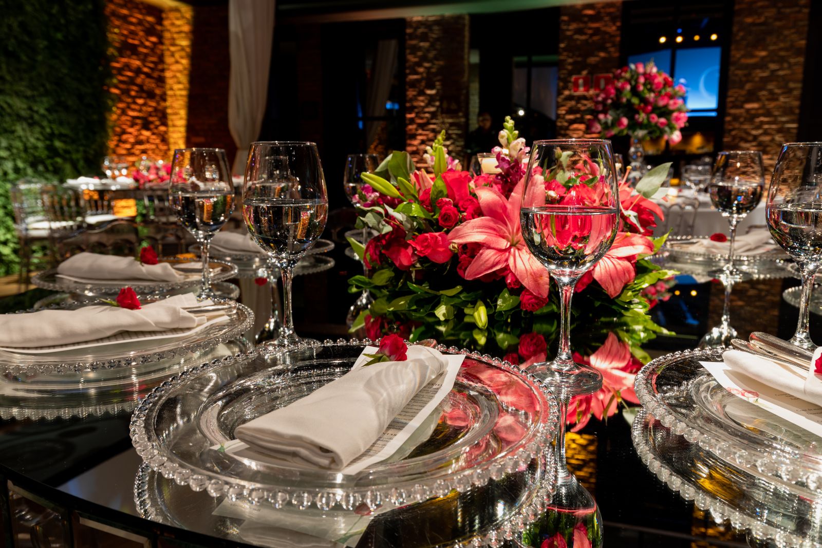 Mesa de banquete de boda con copas llenas de agua, platos de cristal, servilletas blancas y un centro de lirios y rosas rojas