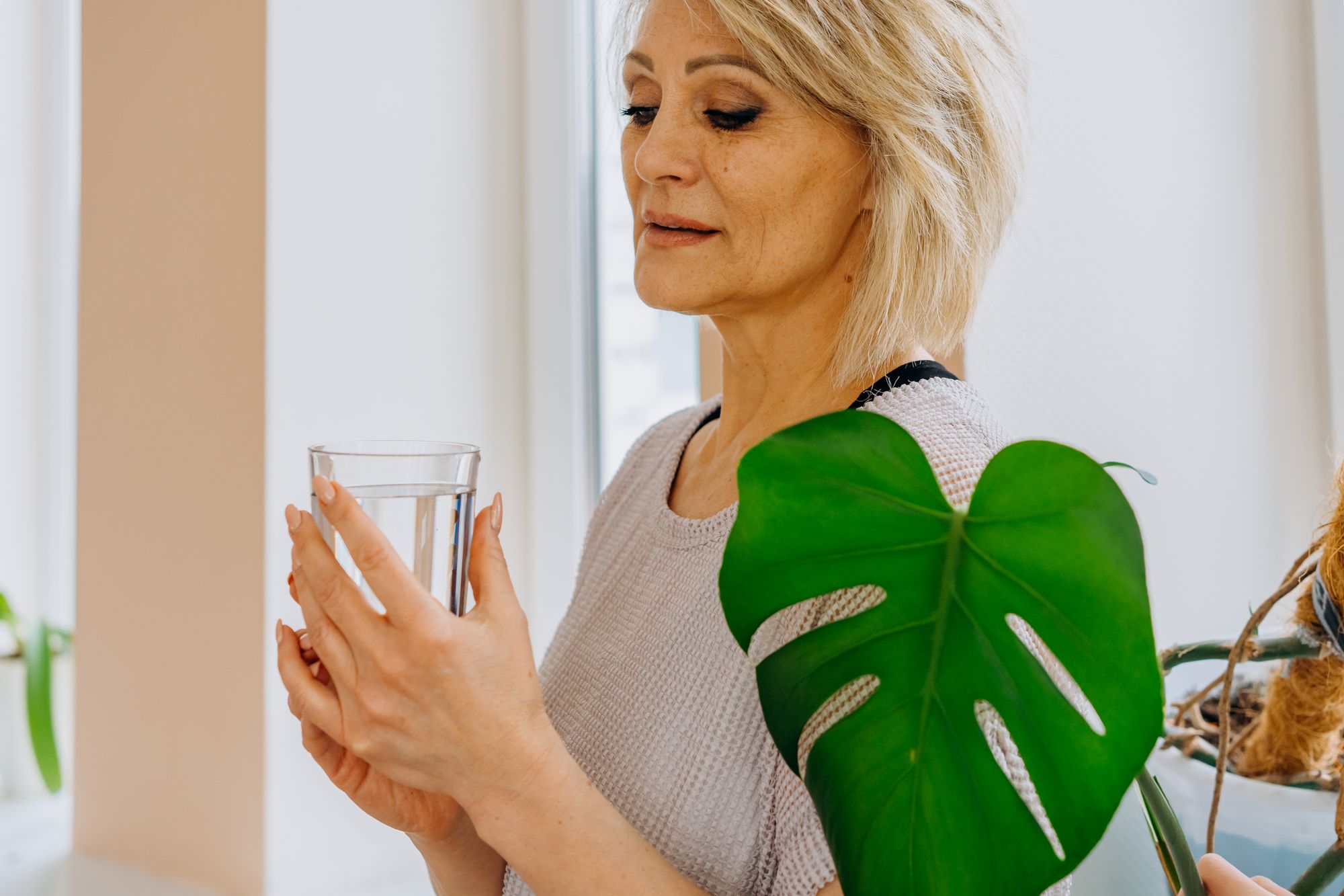 Mujer de mediana edad sosteniendo un vaso de agua con las dos manos junto a una ventana luminosa, en una estancia con plantas