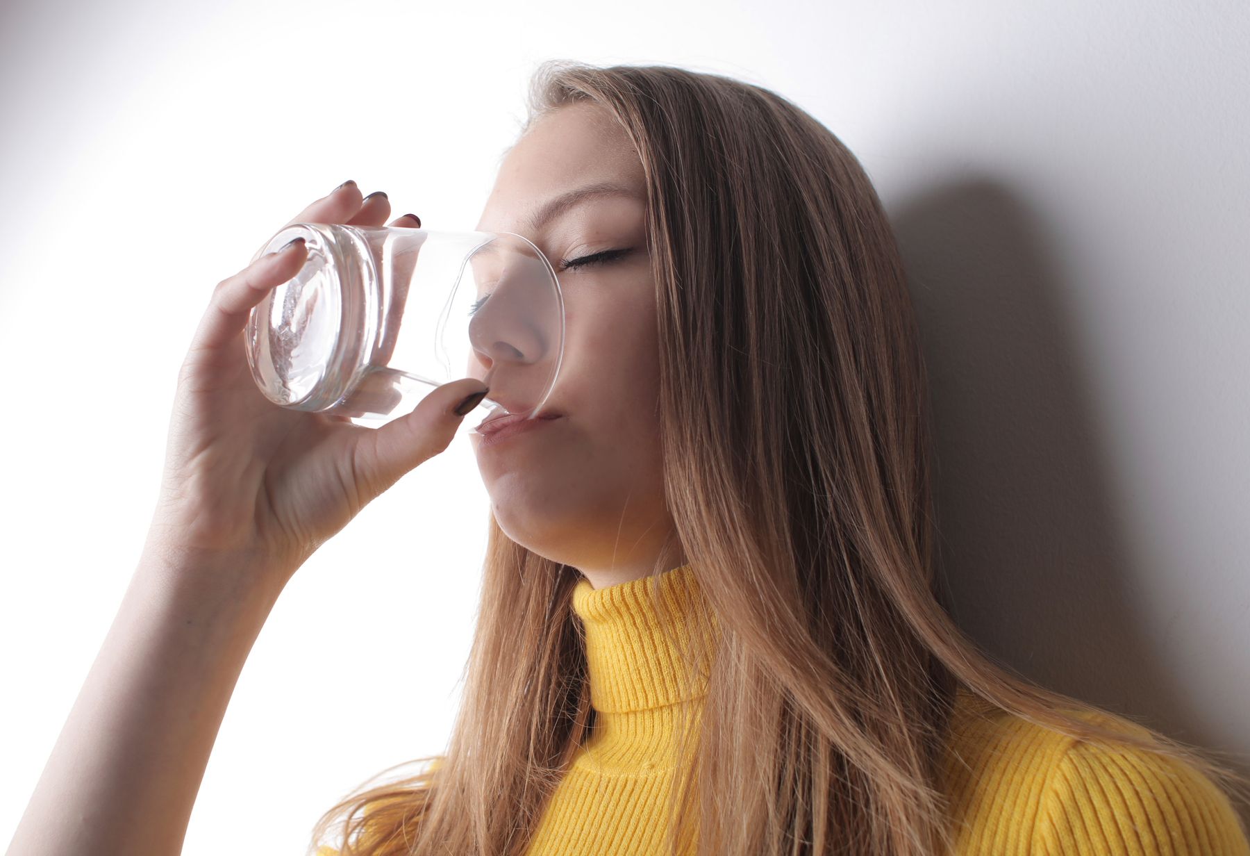 Mujer con jersey de cuello alto amarillo bebiendo un vaso de agua en interior, un gesto que en oto&ntilde;o hay que recordar porque la sed deja de avisar