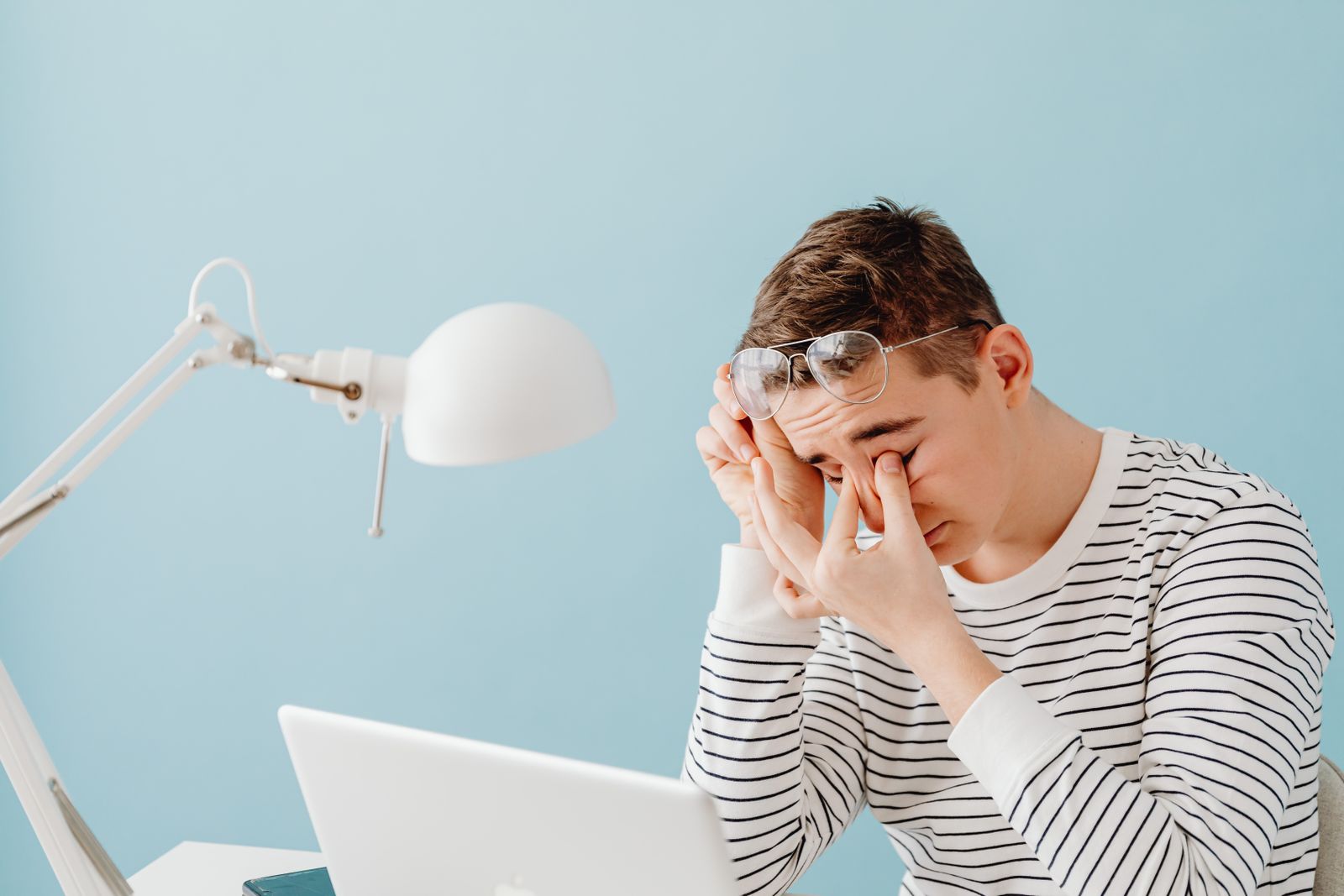 Joven con gafas levantadas frot&aacute;ndose los ojos cansados delante de un port&aacute;til, junto a un flexo blanco sobre fondo azul claro