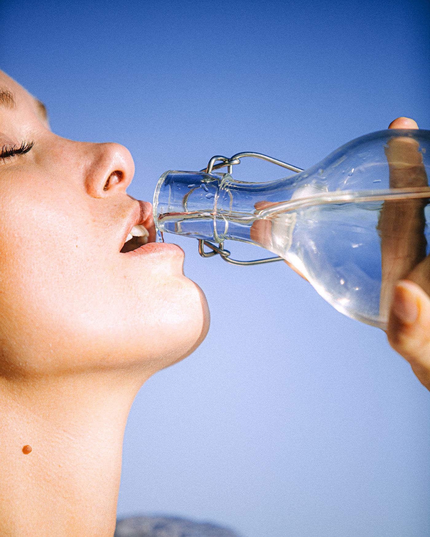 Persona bebiendo agua de una botella de cristal contra un cielo azul para mantener la hidrataci&oacute;n corporal