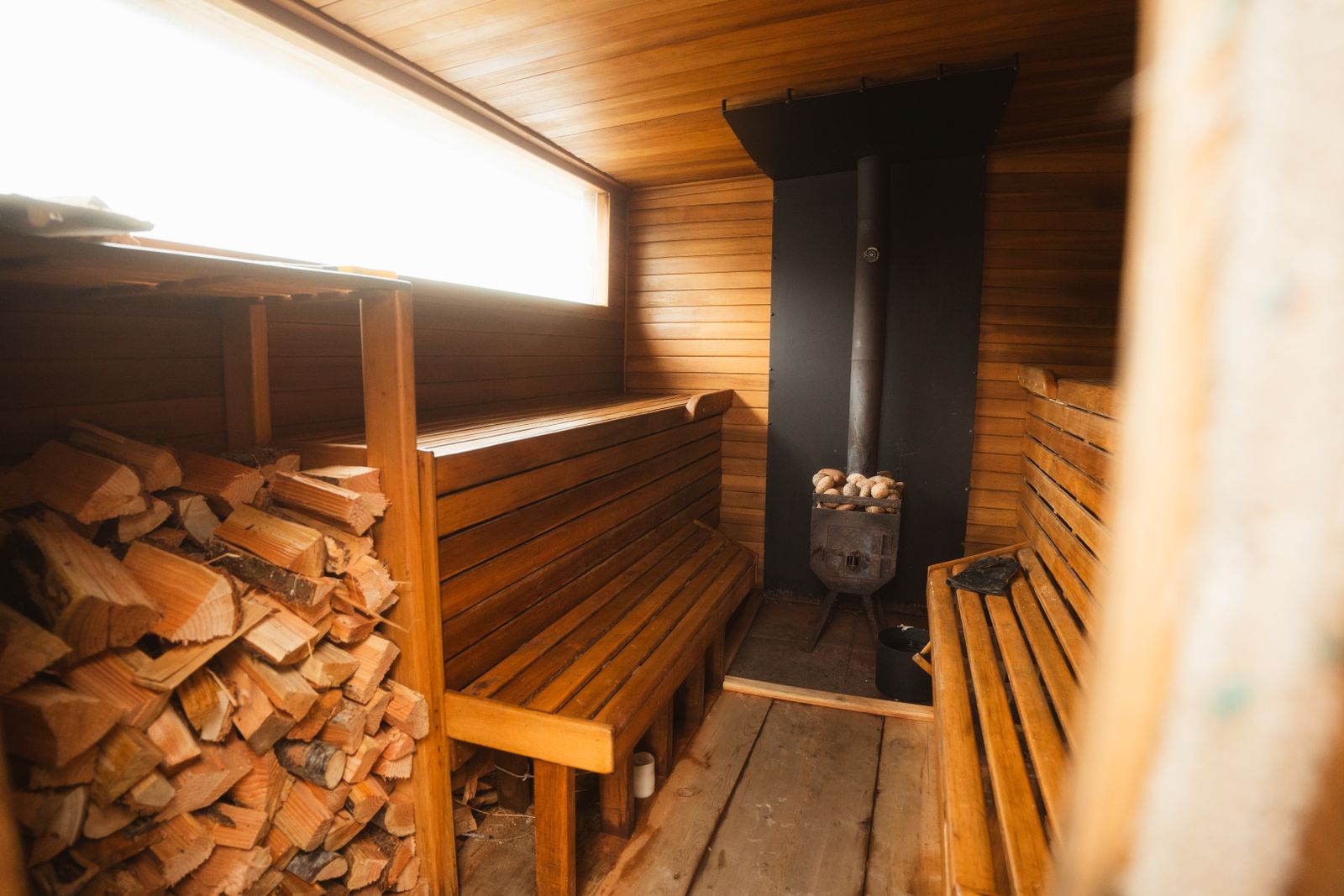 Interior de una sauna finlandesa de madera con estufa de le&ntilde;a, bancos y troncos apilados junto a la ventana