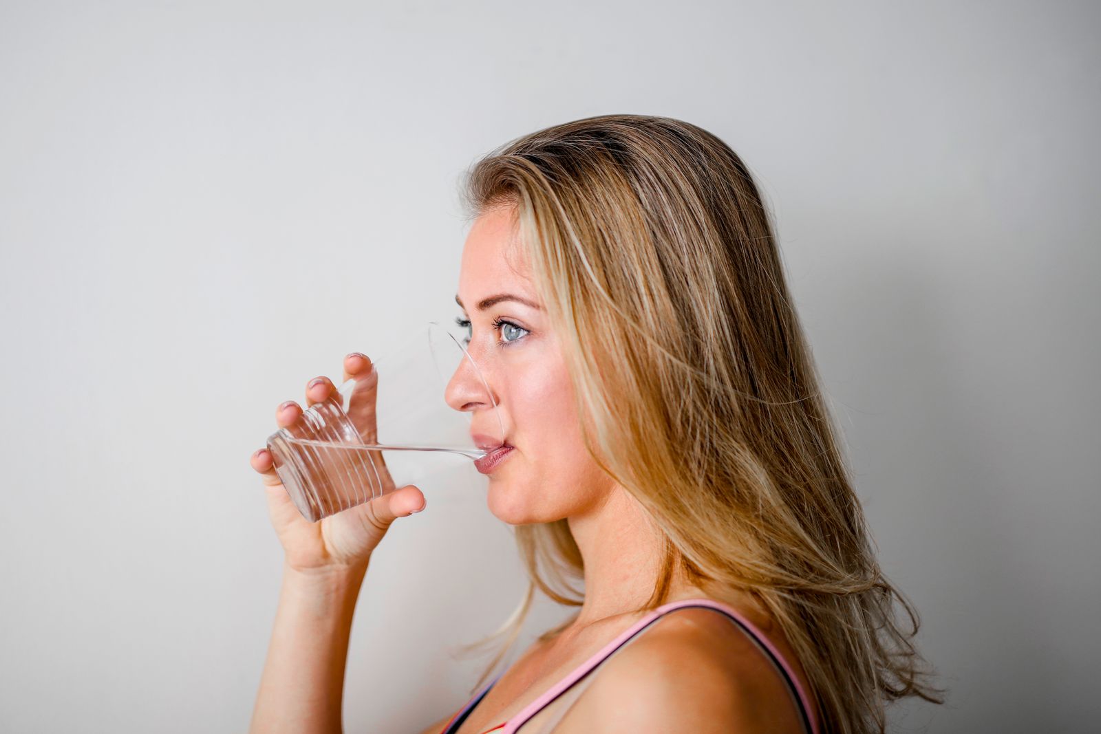 Mujer de perfil bebiendo un vaso de agua frente a una pared clara