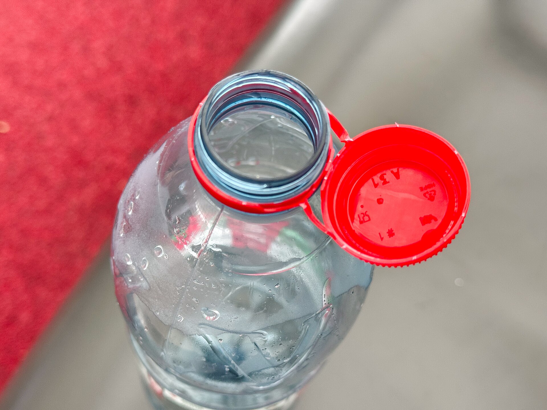 Primer plano del cuello de una botella de agua de pl&aacute;stico abierta con el tap&oacute;n rojo sujeto al anillo por su bisagra, conforme a la normativa europea
